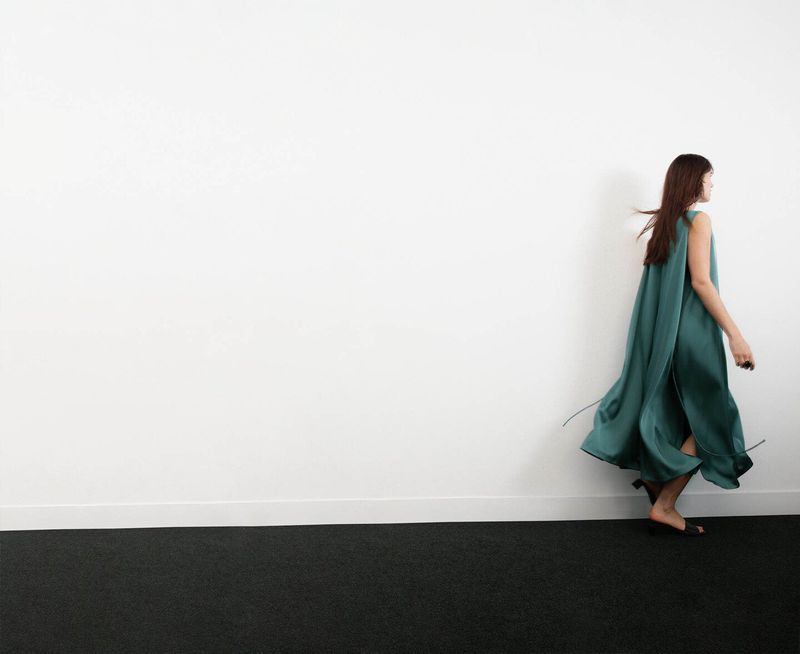 Model in a long flowing green dress walking across a minimalist studio setting with a white wall
