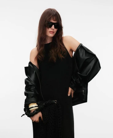 Model in dark sunglasses and black dress wearing a black leather jacket against a white background