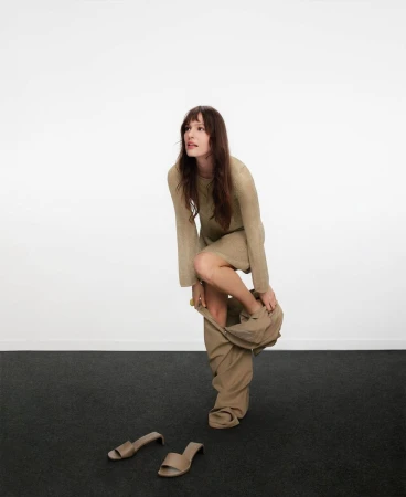 Model in neutral dress stepping out of trousers against a white backdrop with a dark floor