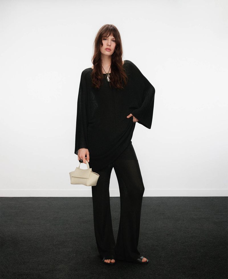 Model standing against a white wall wearing a black tunic and trousers while holding a light handbag