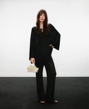 Model standing against a white wall wearing a black tunic and trousers while holding a light handbag