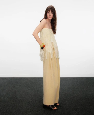 Model wearing a strapless fringe top and wide-leg trousers standing against a plain white studio backdrop with dark carpet.