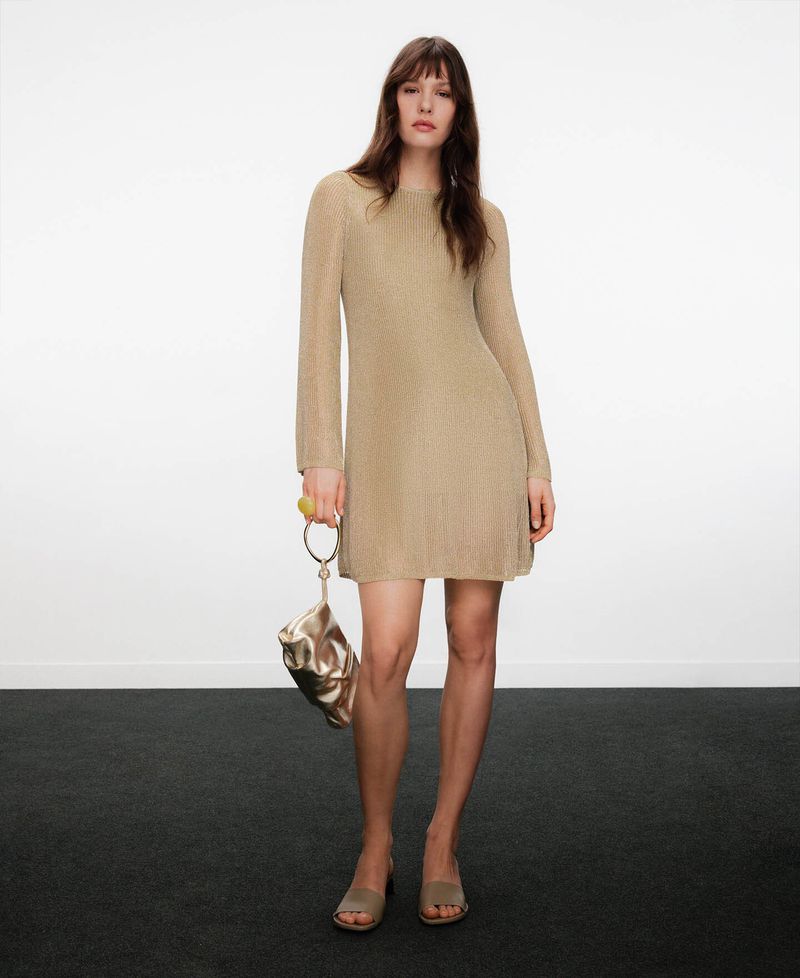 Model wearing a long-sleeved gold knit dress and carrying a metallic gold handbag against a plain background