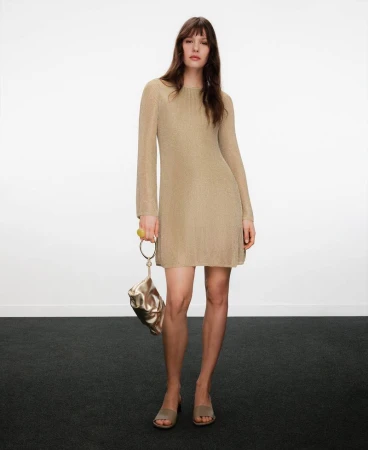 Model wearing a long-sleeved gold knit dress and carrying a metallic gold handbag against a plain background