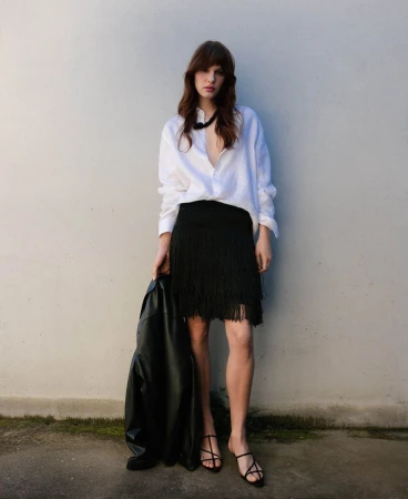 Model wearing a white shirt and black fringed skirt holding a black jacket against a plain wall