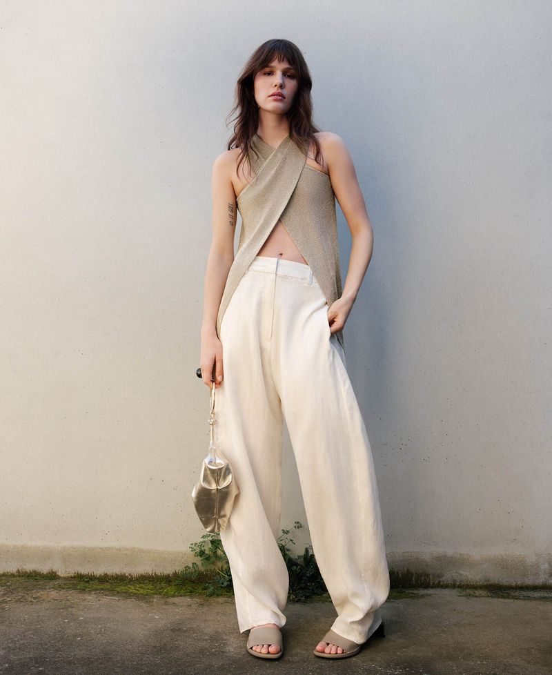 Fashion model in a cross-halter top and loose white pants poses against a neutral wall holding a metallic fish-shaped purse.