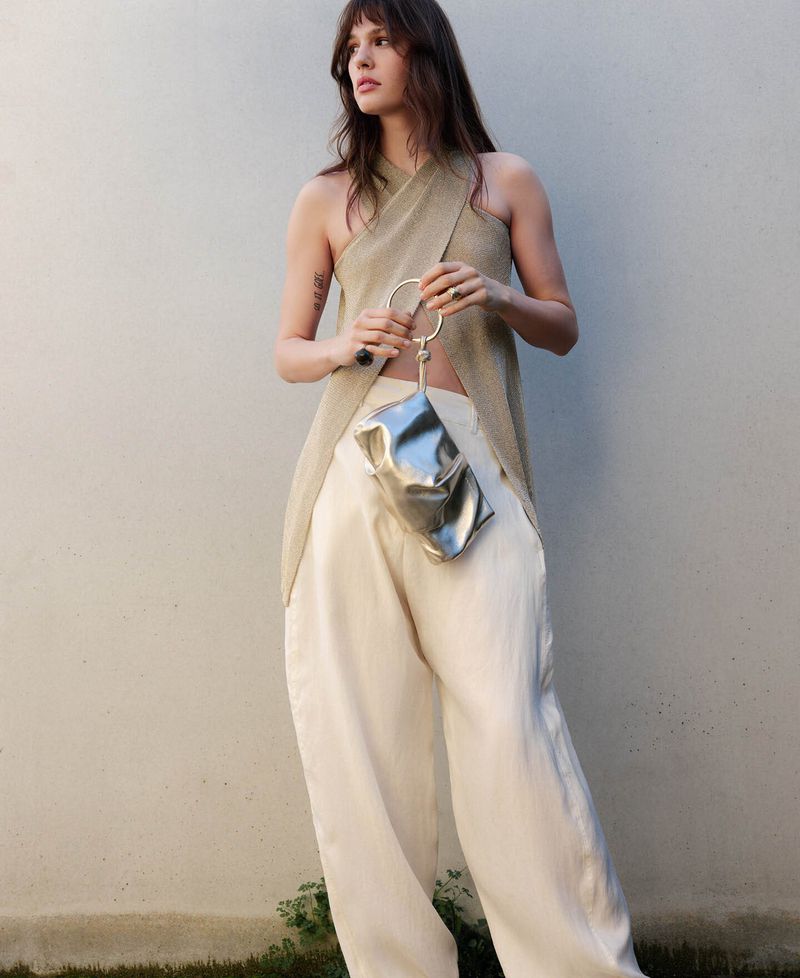 Model wearing a halter top and light trousers holding a metallic silver handbag against a plain wall for Adolfo Dominguez.