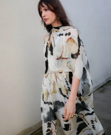 Model wearing a printed watercolor dress with flowing cape-like sleeves against an outdoor wall.