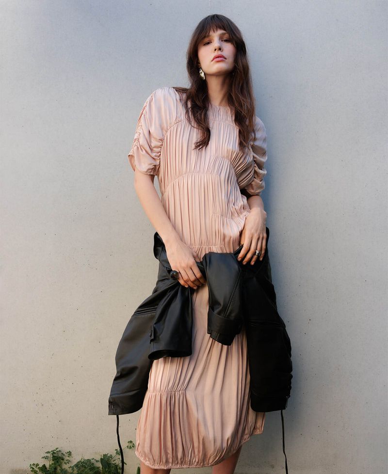 Model in a pink pleated dress holding a black jacket against a plain wall