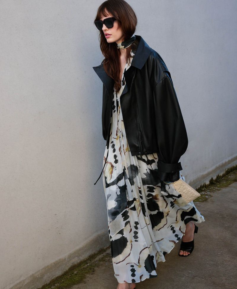 Model wearing a black leather jacket over an abstract print dress and dark sunglasses outdoors against a pale wall.