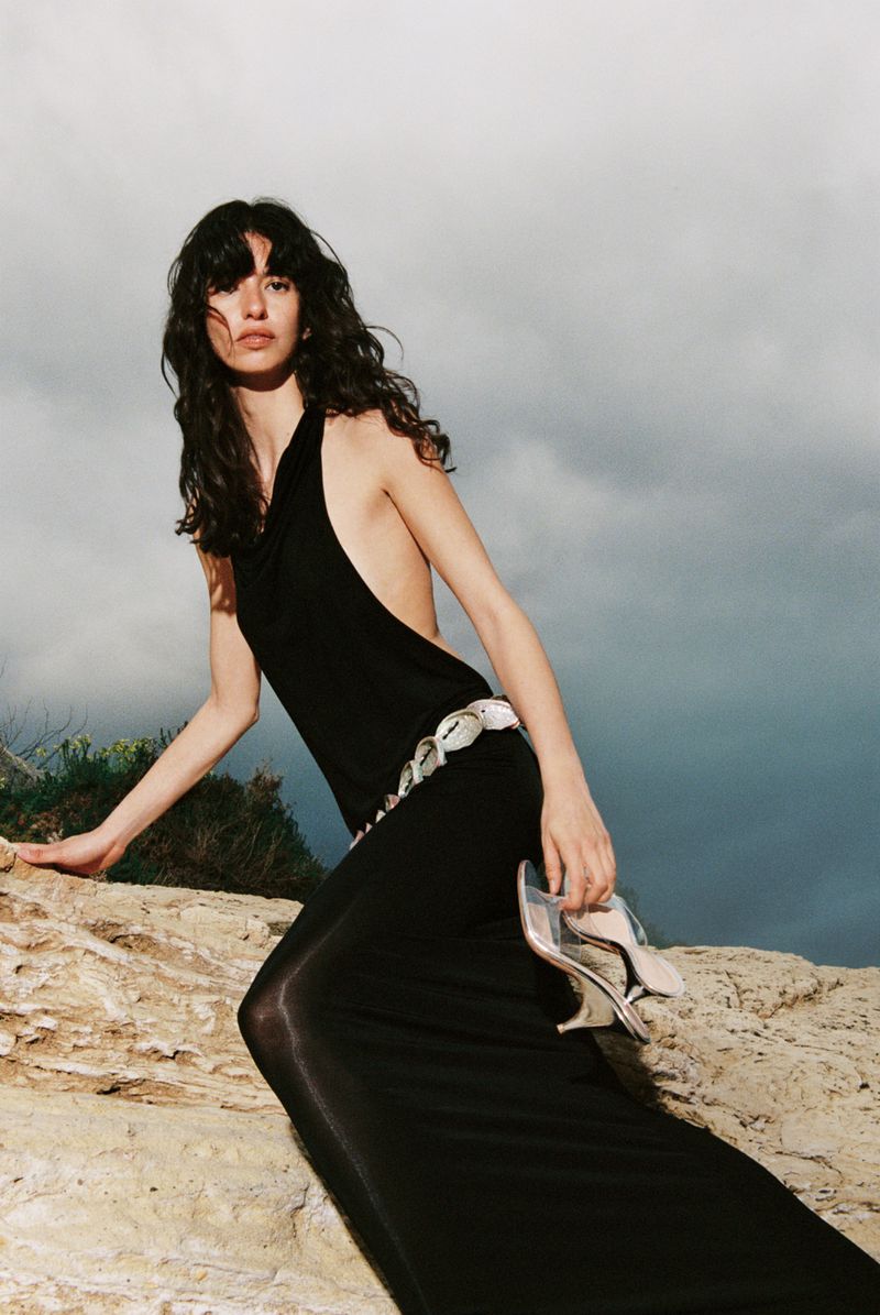 Model in a black maxi dress holding sandals, posed on rocky terrain against an overcast sky.
