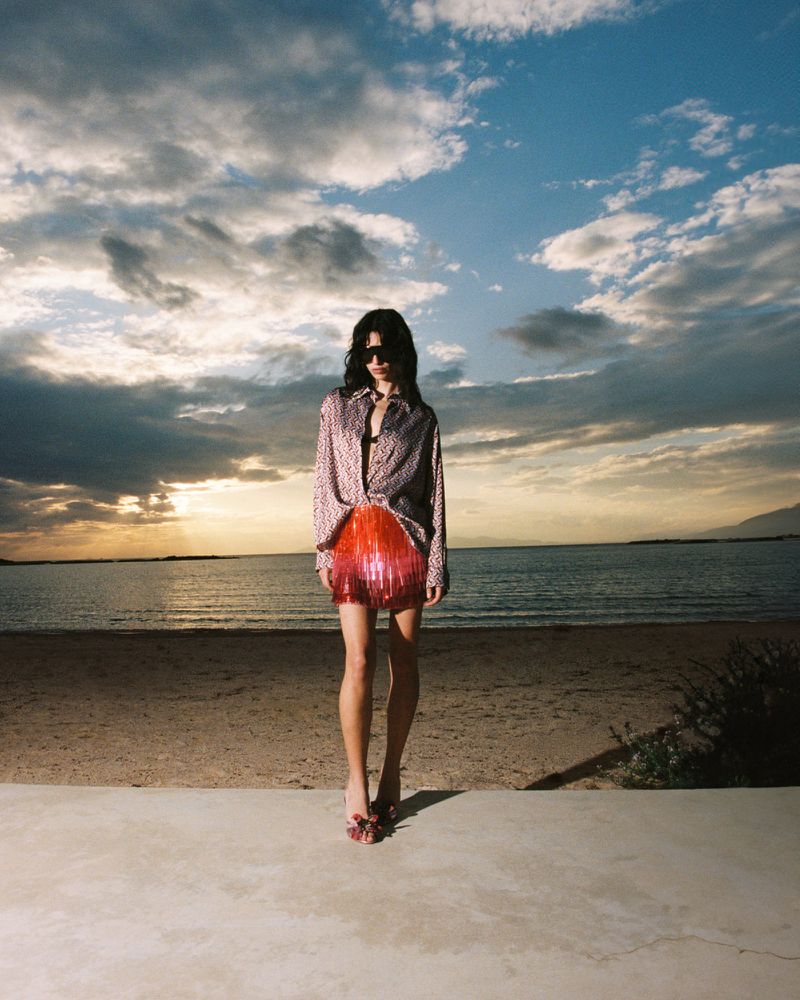 Model wearing patterned shirt and red fringed skirt standing on beach at sunset