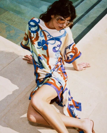 Model wearing a patterned silk dress and tinted sunglasses seated by a concrete pool edge for Moda Operandi.