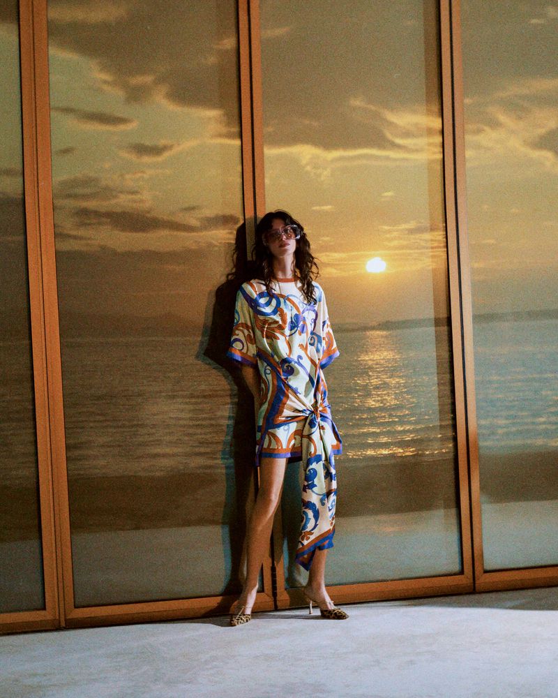 Model wearing patterned silk dress leaning against glass door reflecting sunset over ocean for Moda Operandi