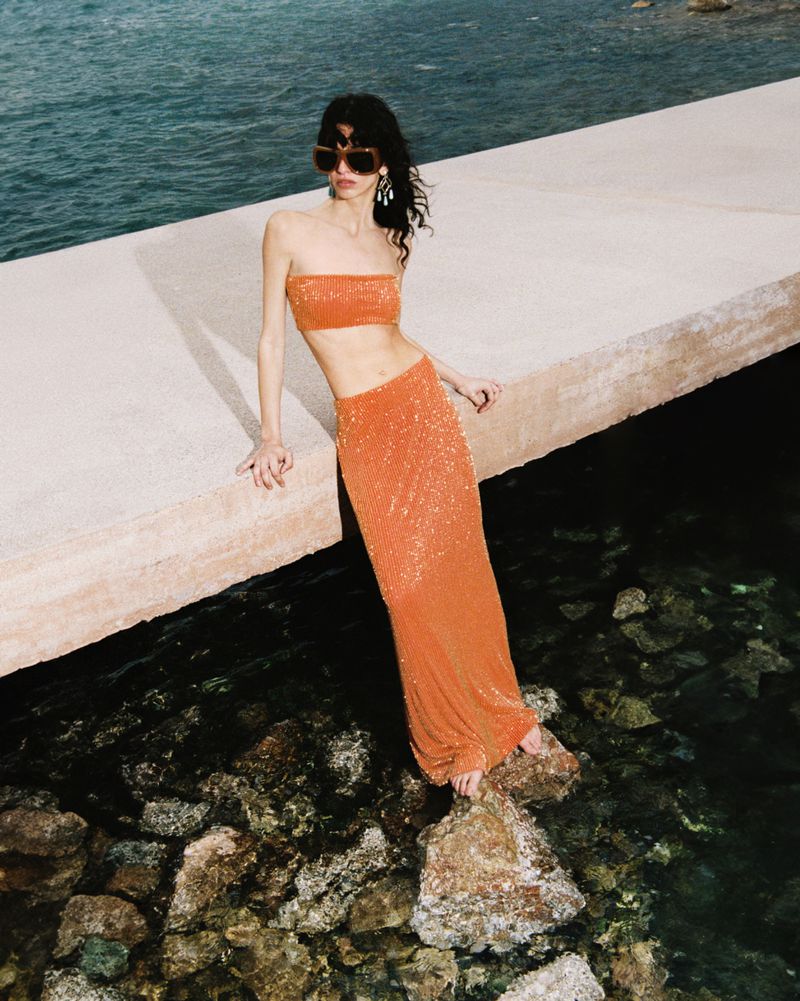 Model leaning on a concrete pier in an orange two-piece outfit with sunglasses above dark water.