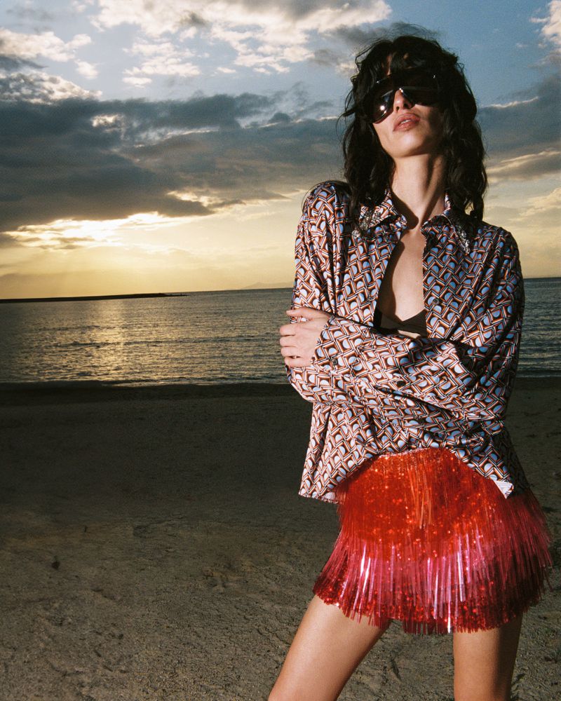A woman poses on a beach in a patterned shirt and orange skirt with the ocean and a colorful sunset sky in the background.