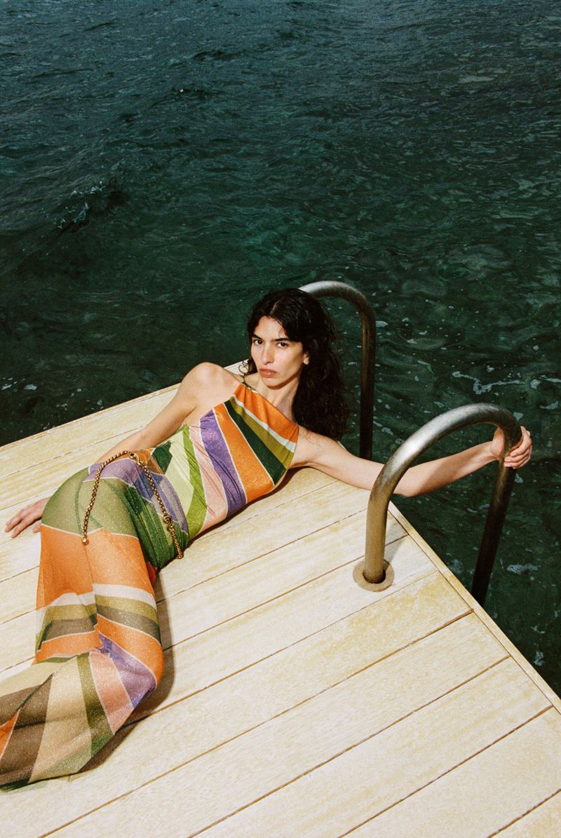 Model in a striped halterneck dress lying on a wooden deck next to pool ladder rails by dark water for Club Moda La Isla