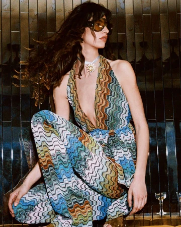 Model wearing a multi-coloured chevron pattern halterneck jumpsuit and tinted sunglasses against a dark panelled wall