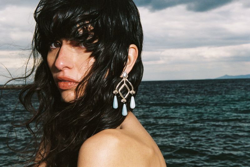 Close-up portrait of a woman with wet dark hair wearing ornate dangling earrings against an ocean and cloudy sky backdrop.