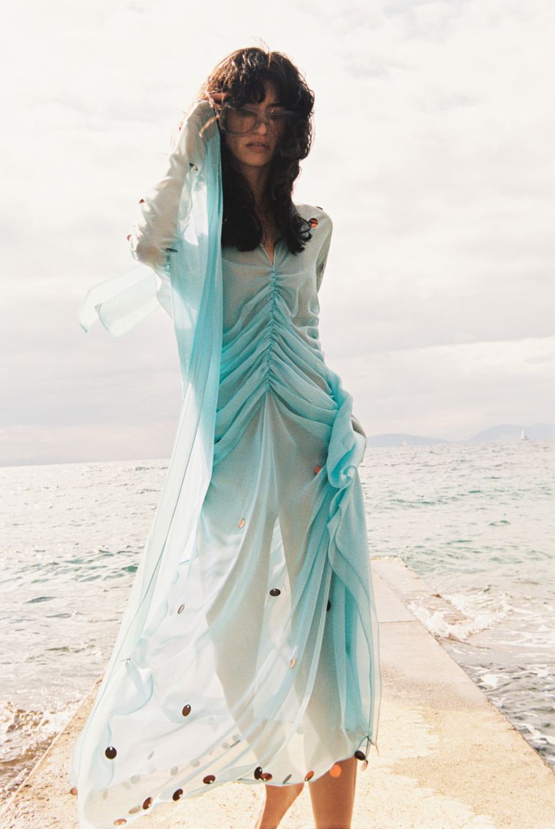 Model in a light blue sheer dress posing on a concrete pier by the ocean under an overcast sky.