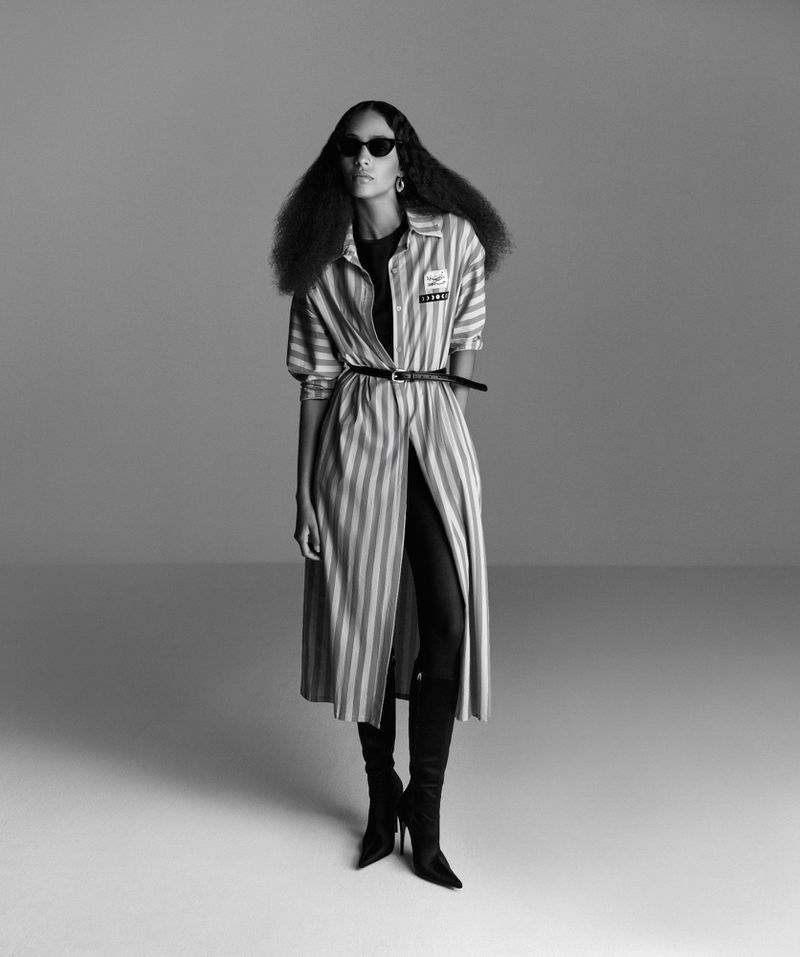 Black and white fashion shot of a black female model in a striped coat, black sunglasses, and boots posing in a studio.