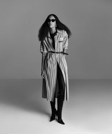 Black and white fashion shot of a black female model in a striped coat, black sunglasses, and boots posing in a studio.