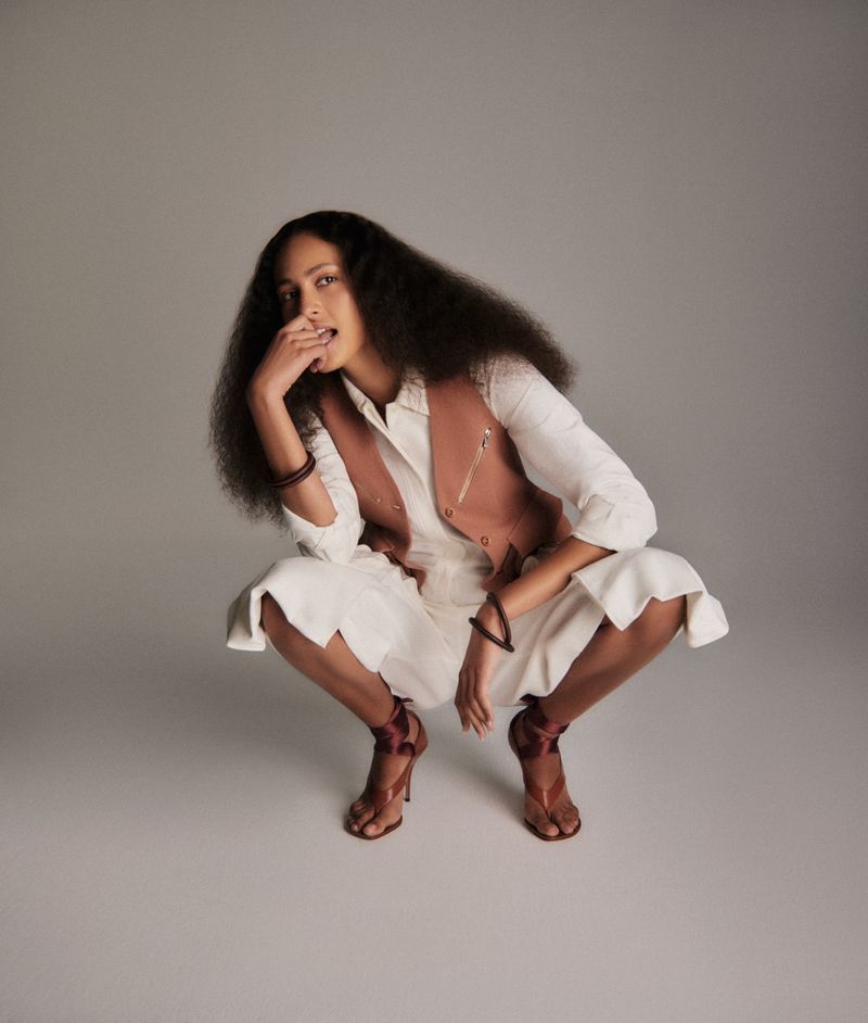 Fashionable woman with curly hair poses in a crouch, wearing a white linen suit with a brown vest and high-heeled, open-toed shoes.