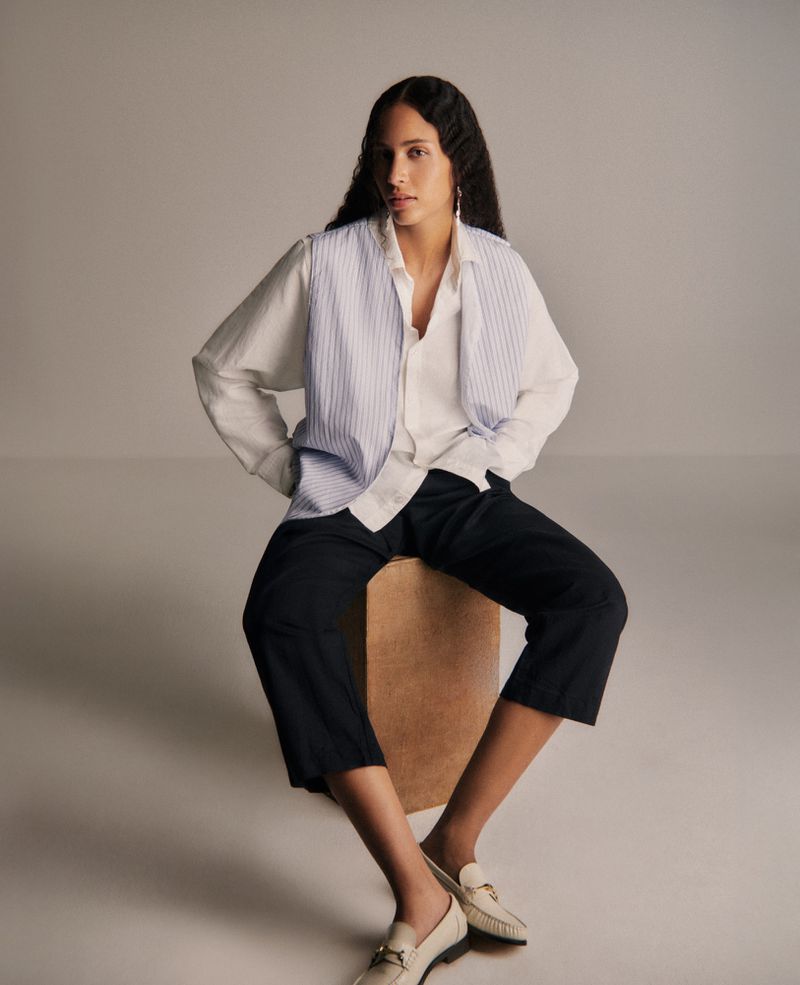 A woman poses while sitting on a box, wearing a white blouse, blue vest, black pants, and white loafers in a neutral studio setting.