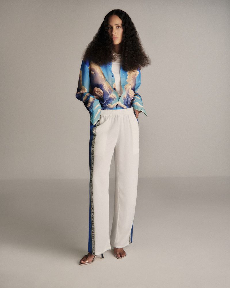 Fashion shot of a woman wearing a blue cloud-print shirt and white wide-leg pants standing in a neutral-toned photo studio.