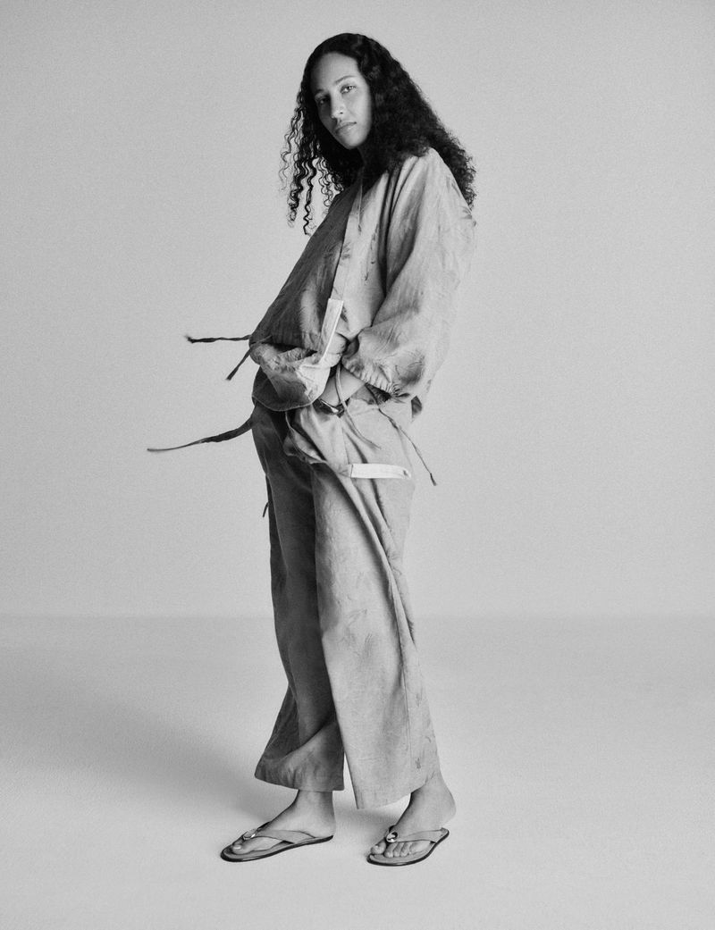 Black and white portrait of a young woman with curly hair, wearing a long-sleeved jacket and pants in a light material, posing in a studio.