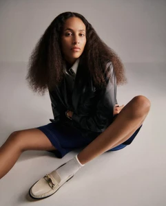 Model seated on a white seamless background wearing a dark leather blazer, blue shorts, white socks, and cream loafers.