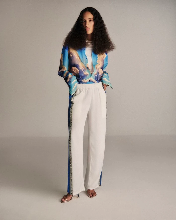Model with long dark curly hair wearing a blue sky-print shirt and white trousers against a neutral studio backdrop