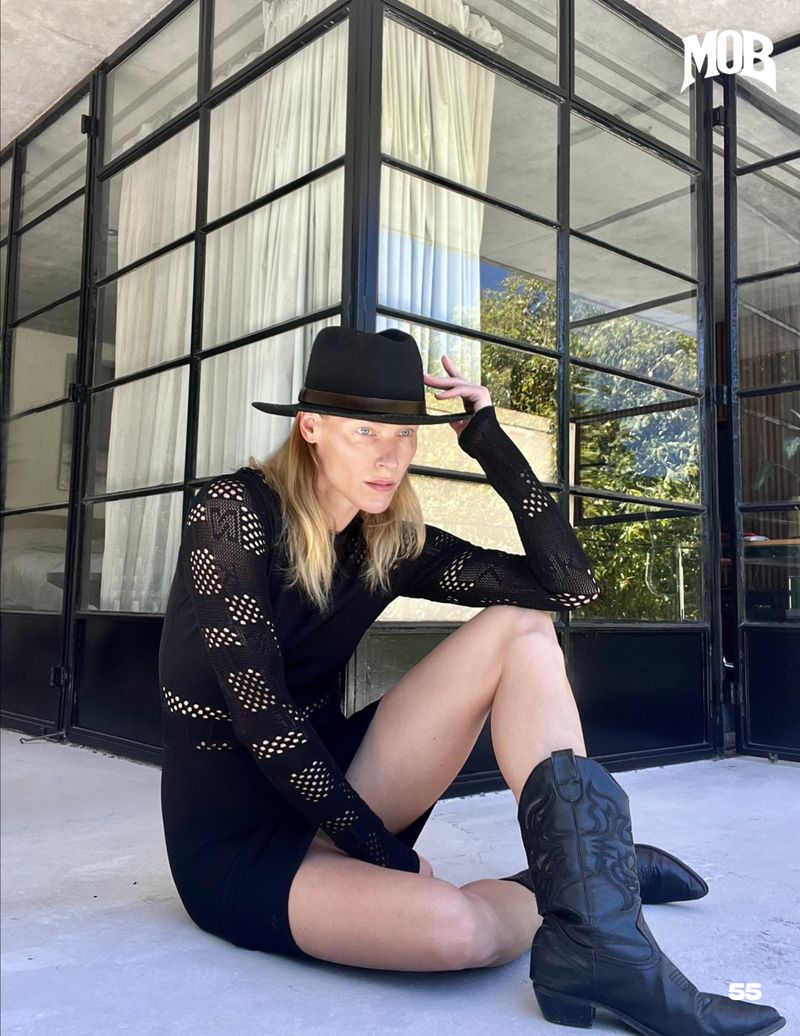 An adult female is sitting on a patio with cowboy boots and a hat, posing in front of a windowed wall, possibly for a fashion editorial.