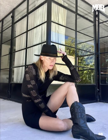 An adult female is sitting on a patio with cowboy boots and a hat, posing in front of a windowed wall, possibly for a fashion editorial.