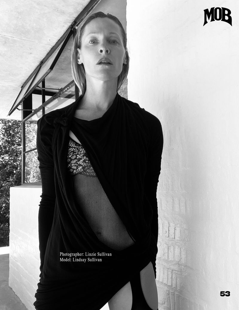 Black and white portrait of a model in a black dress, lace top and dramatic draping, posing against a wall with architectural detail.
