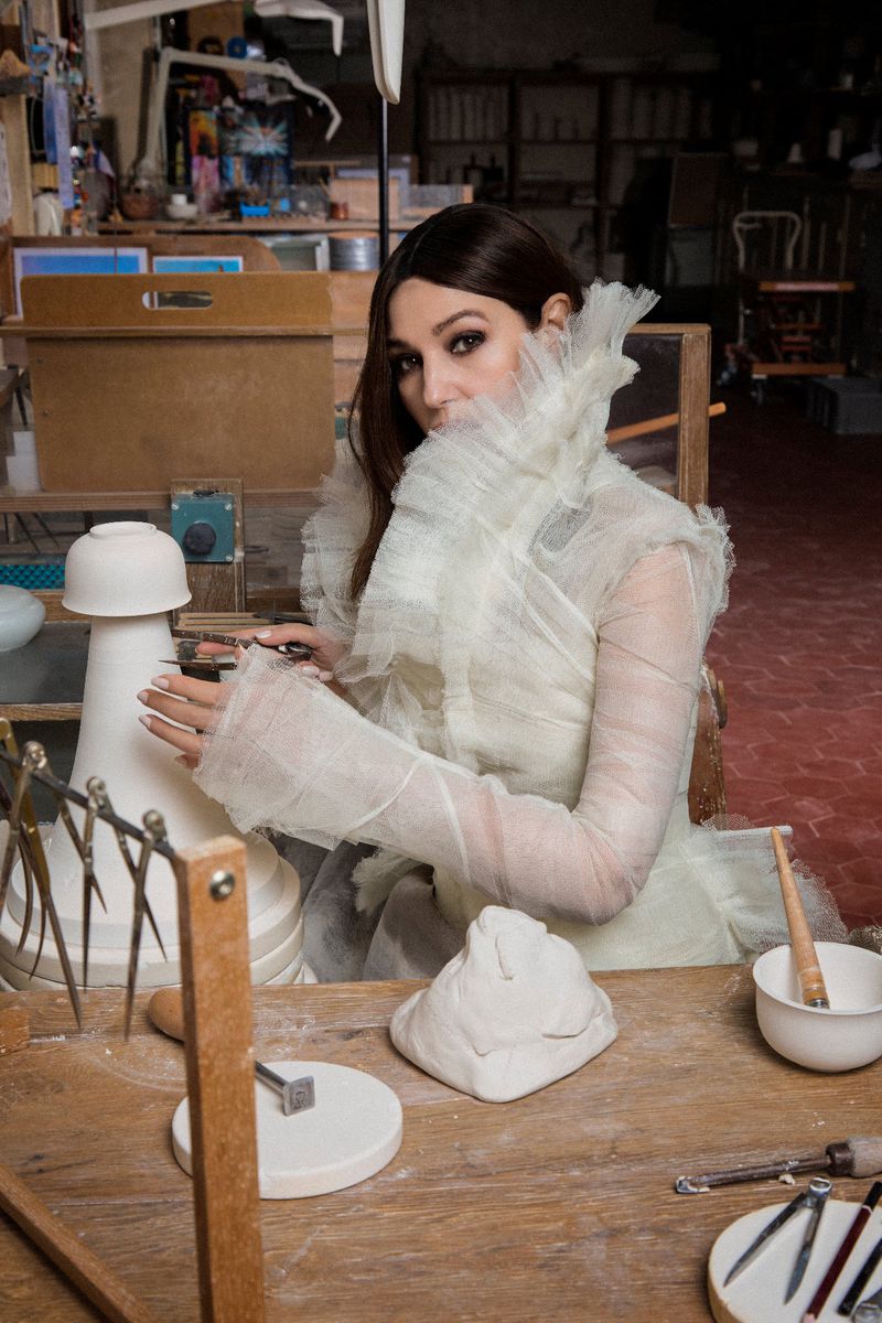Woman in ruffled collar and long sleeves sits at a wooden table with clay, tools, and a partially made pottery lamp.