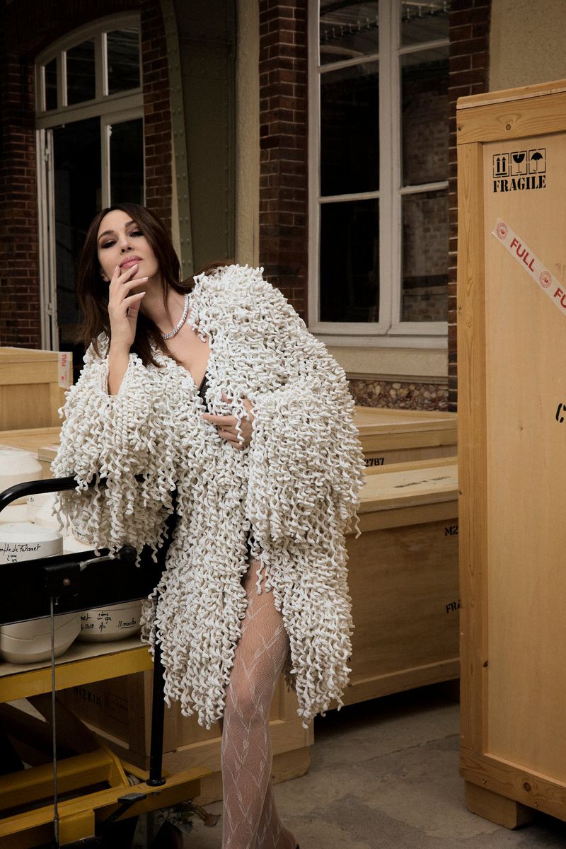 Adult woman posing in a fringed white coat with interior brick wall and wooden crates in the background. Fashion or editorial shot.
