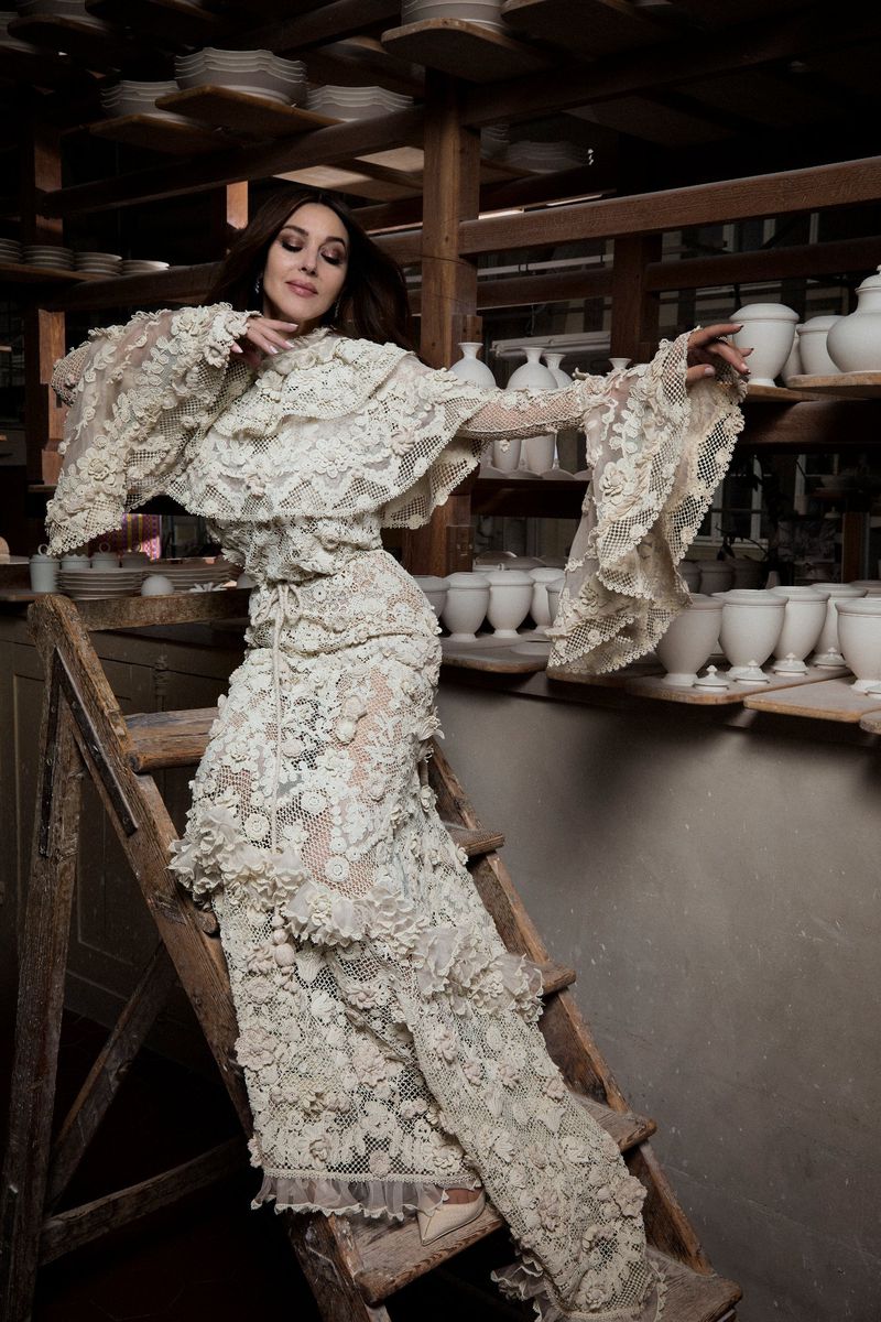 Monica Bellucci poses on wooden ladder in a long, ivory-colored, floral lace dress in front of shelves of pottery in a studio.