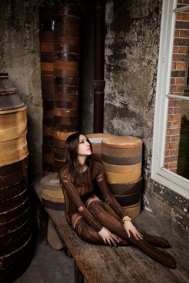 Asian woman in brown dress and thigh-high boots posing on a wooden bench. Brown and beige cylinders in the background, natural light.