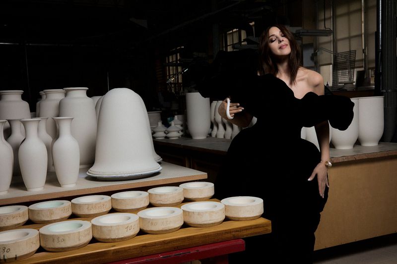 Monica Bellucci in a black couture dress poses in front of vases and ceramic pieces in a factory setting with dark and moody lighting.