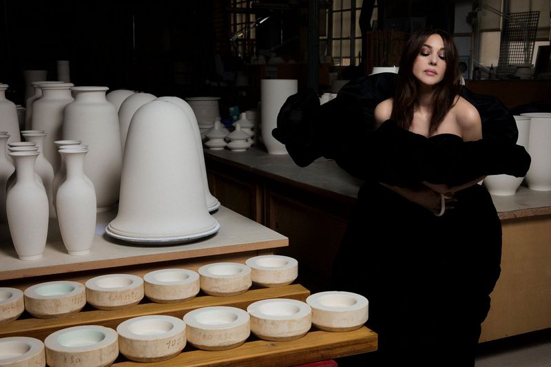 Monica Bellucci in a black dress poses near pottery and a wooden table in an indoor setting with soft lighting and shallow depth of field.