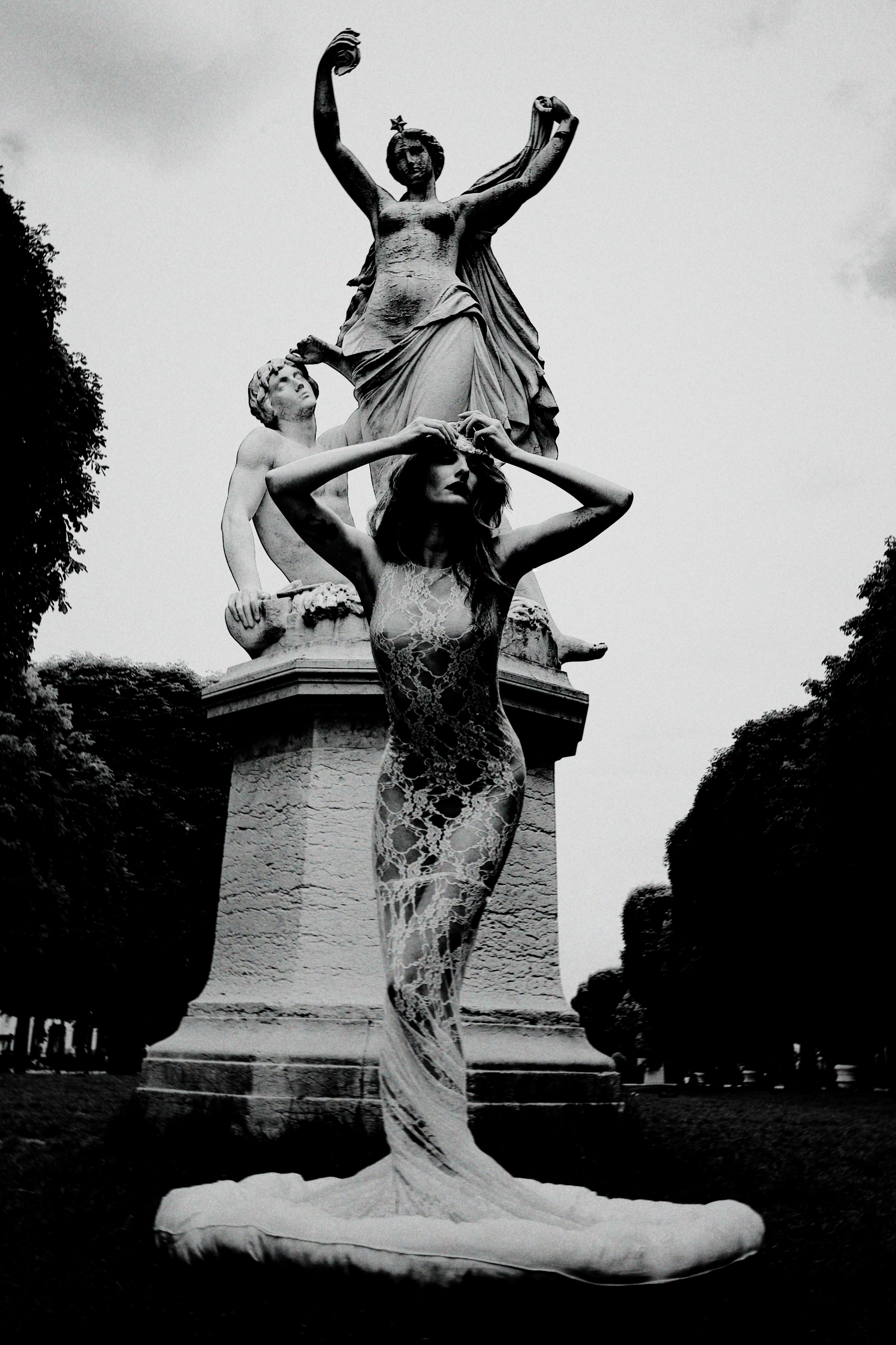 A fashion model in a sheer gown poses dramatically in front of a classical stone statue in an outdoor park.