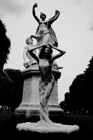 A fashion model in a sheer gown poses dramatically in front of a classical stone statue in an outdoor park.