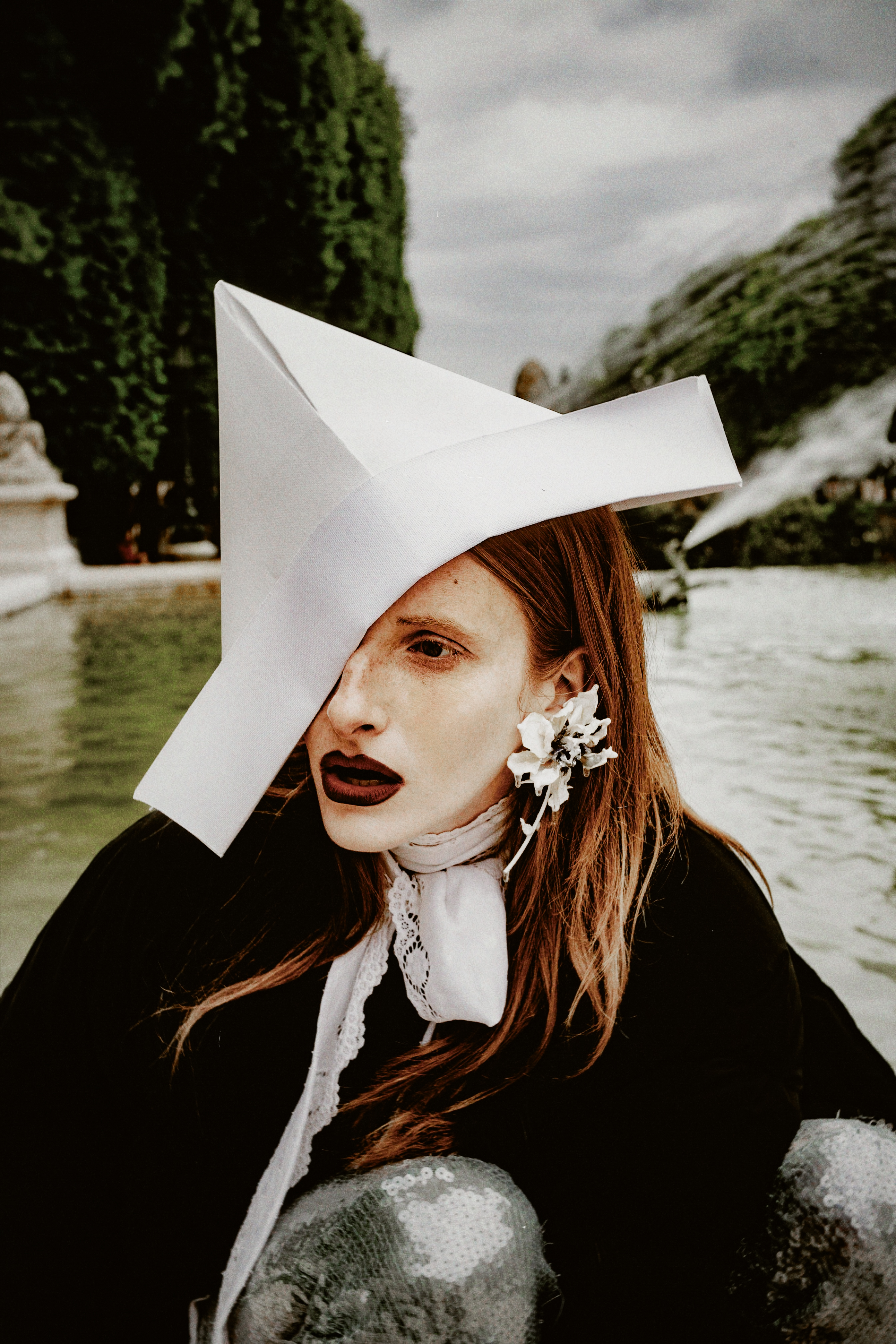 A woman wearing a large white hat and dark clothing poses by a fountain with green foliage in the background.