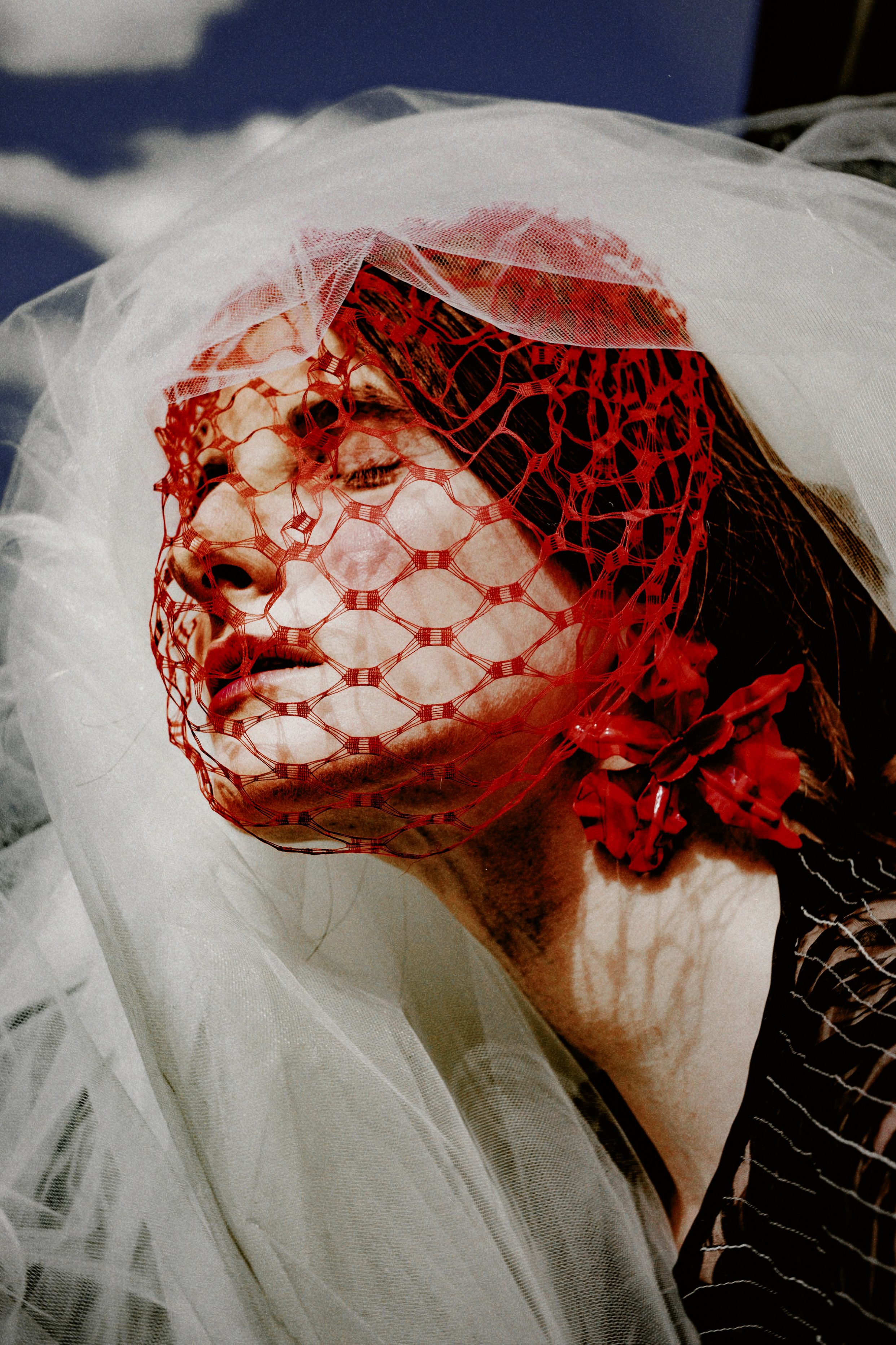 Close-up of a model wearing a red netted veil and white tulle headpiece against a cloudy sky
