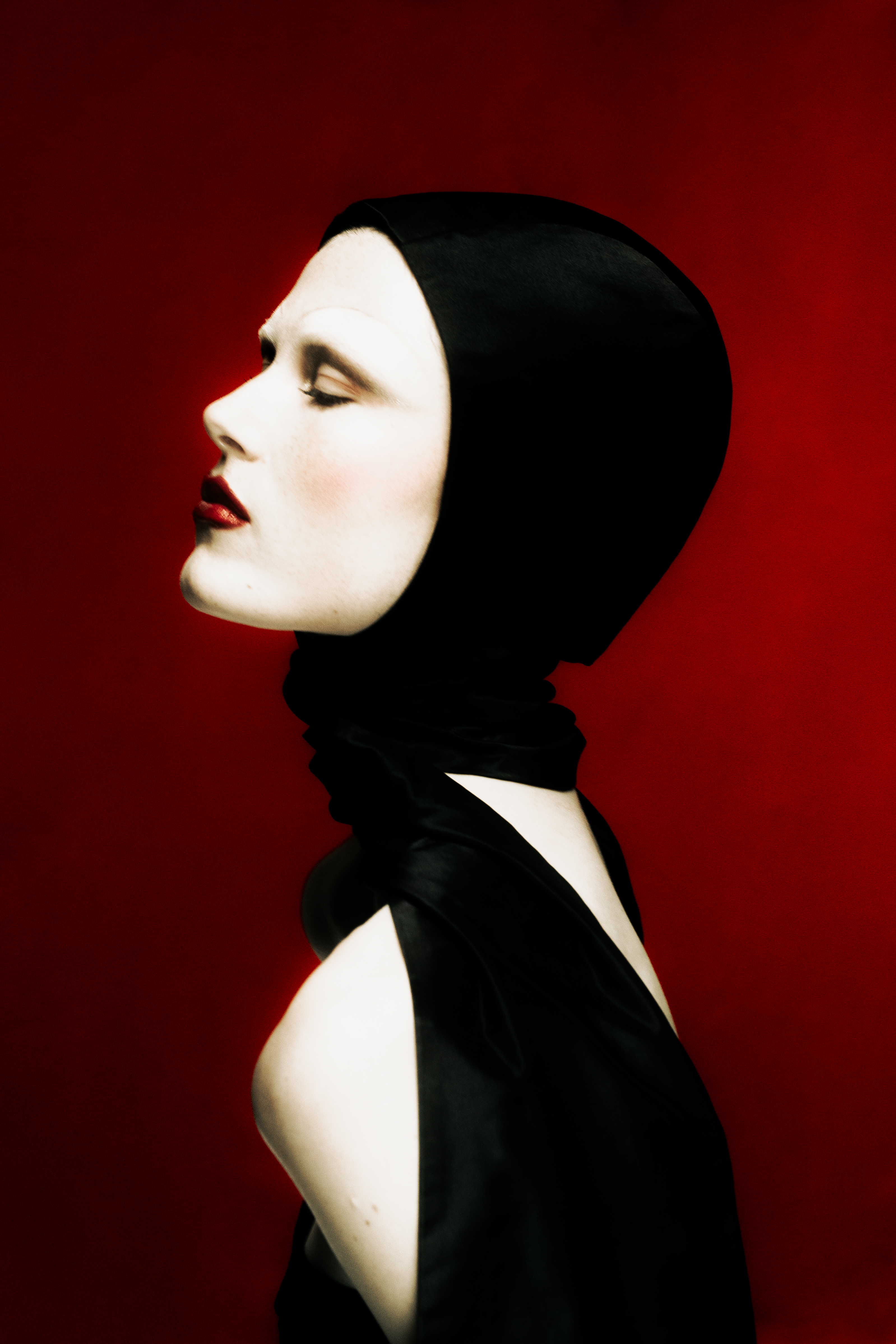 A person in a black head covering and garment with pale skin against a striking red background in a profile view.