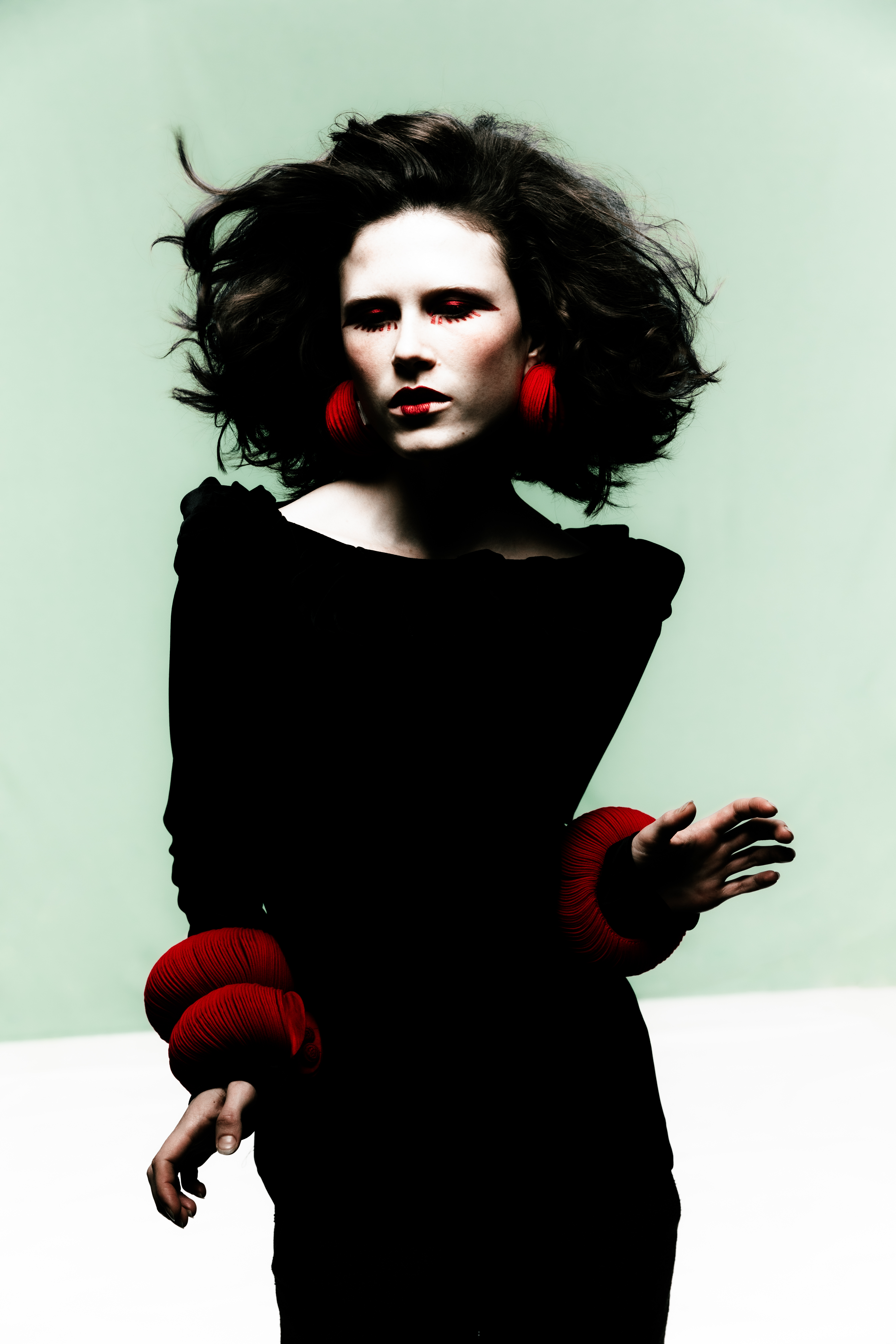 Model with voluminous dark hair wearing a black top and bold red accessories against a plain background