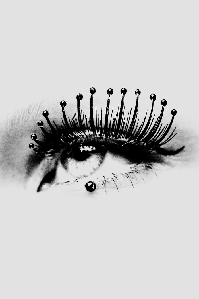 Close-up macro shot of a human eye featuring dark eyelashes adorned with small black pin-like spherical beads.