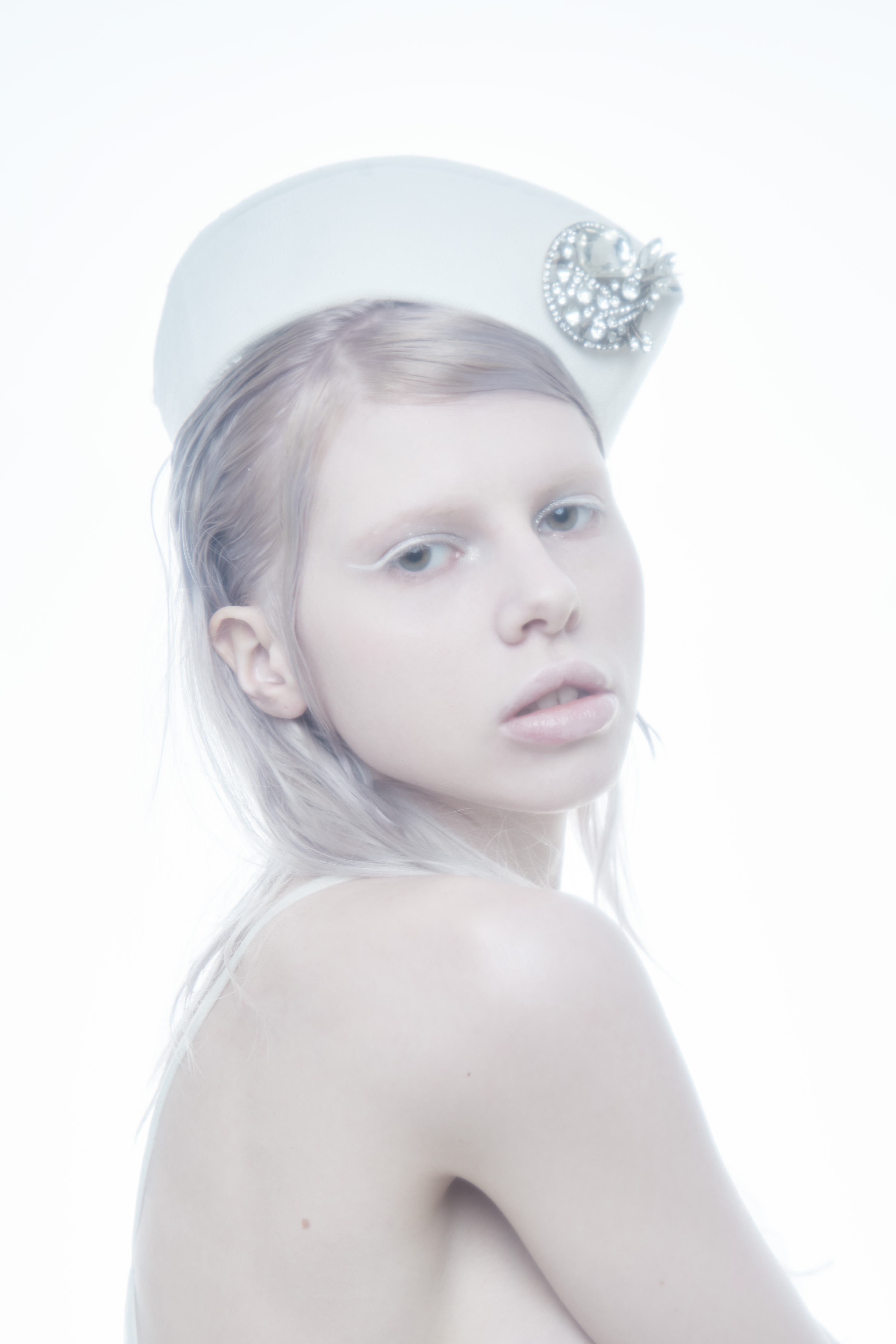 Model wearing a white pillbox hat adorned with crystal brooch against a bright, high-key white seamless backdrop for Flanelle Magazine.