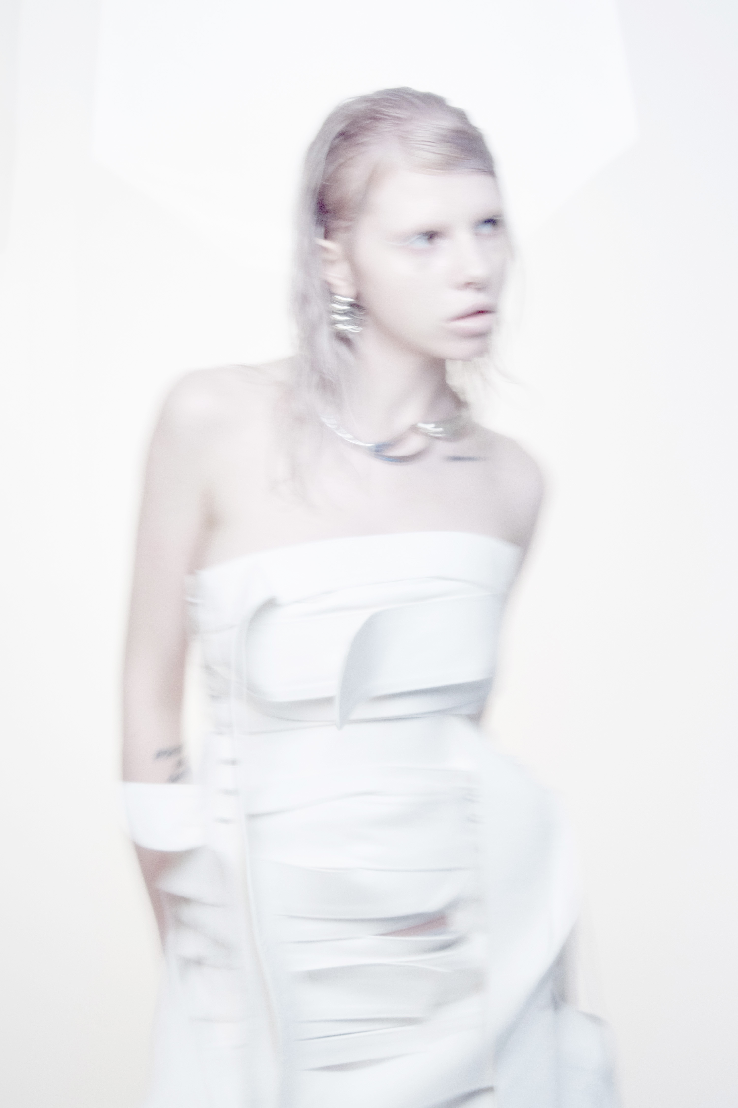 Model with blonde hair and pale styling wearing a structured white strapless dress against a bright white background.